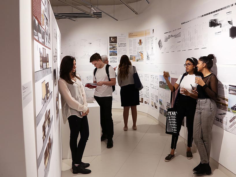 Two students on the far right taking a photo of a friend on the left with people in the background looking at exhibit displays