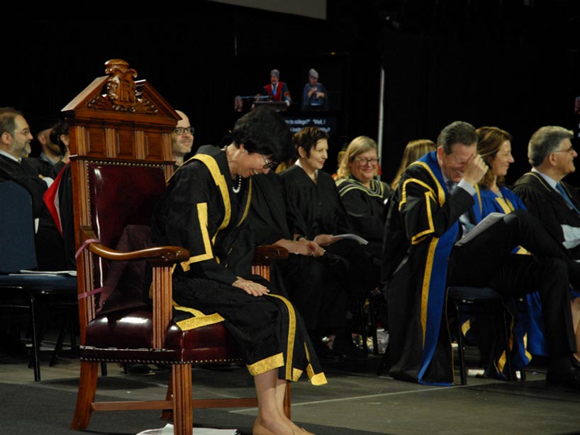 Janice Fukakusa sitting in a wooden chair on stage, laughing, with faculty seated in the background, all wearing convocation gowns