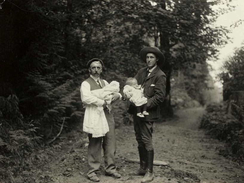 Black and white image of two men in early 1900s suits, standing on a tree-lined path, facing the camera, each carrying a small child