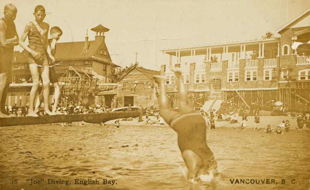 A photograph taken in 1905 depicts lifeguard Joe Fortes diving into the water at English Bay in Vancouver’s Stanley Park, surrounded by people in bathing suits on the dock and on the beach in the background