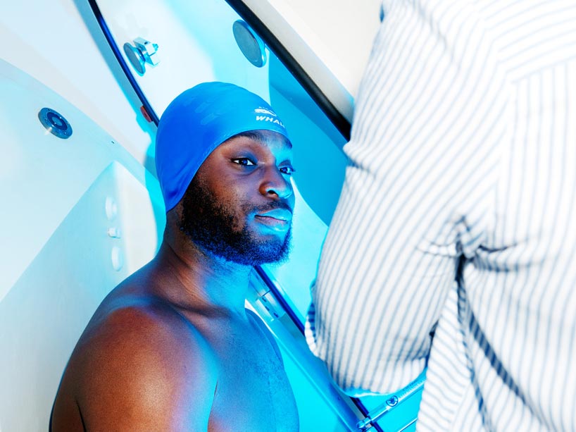 A shirtless man wearing a swimmer’s cap sitting in a pod that assesses body composition