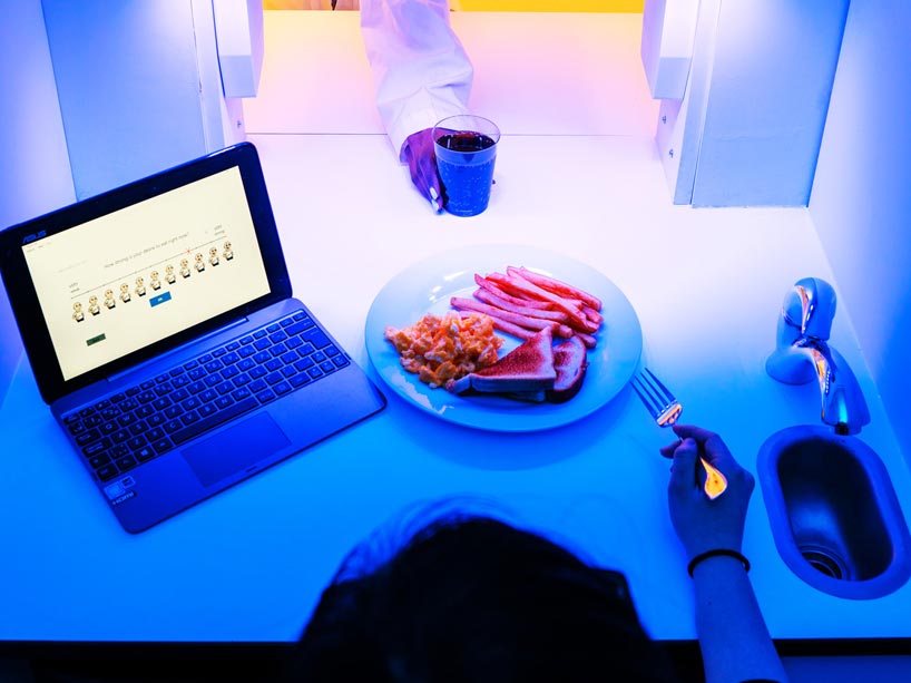 A woman eats a plate of food in a lab while a researcher places a glass in front of her