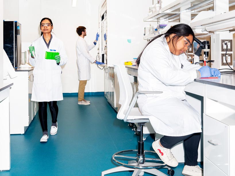 Student carries lab material, another student sits at table looking in microscope and a third student stands near the back of the lab