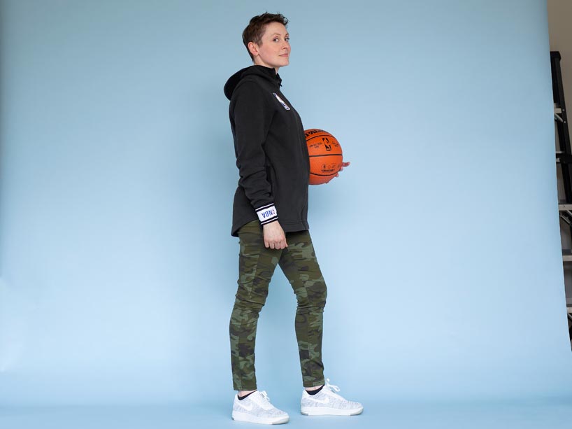 Leah MacNab standing in front of a blue background, facing the camera sideways, holding a basketball in her left hand