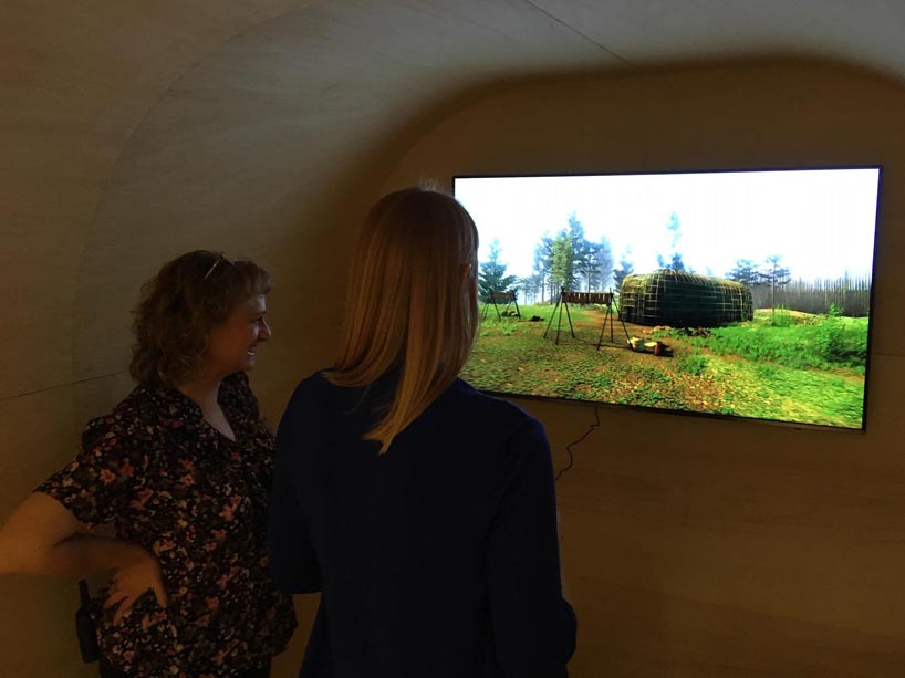 Two women use a controller and TV to view the longhouse