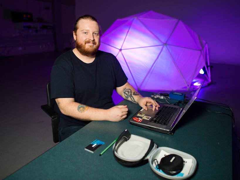 Ryerson grad Aaron Labbé sits at a desk with a laptop computer, smiling at the camera, with a small dome behind him that is equipped with his AI technology
