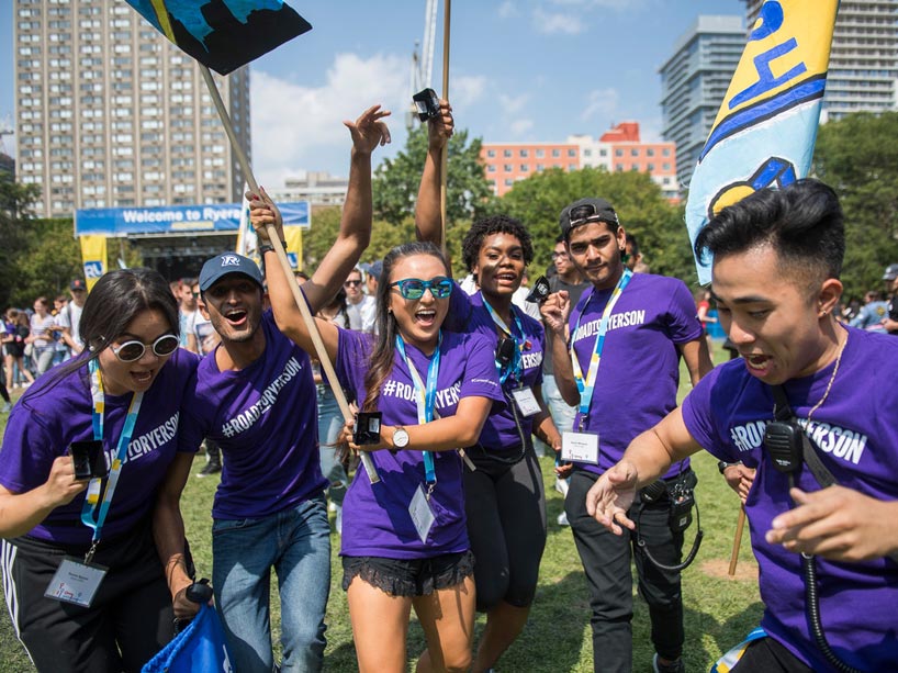 Six students wearing purple t-shirts that say Road to Ryerson holding flags and cowbells, dancing in the quad