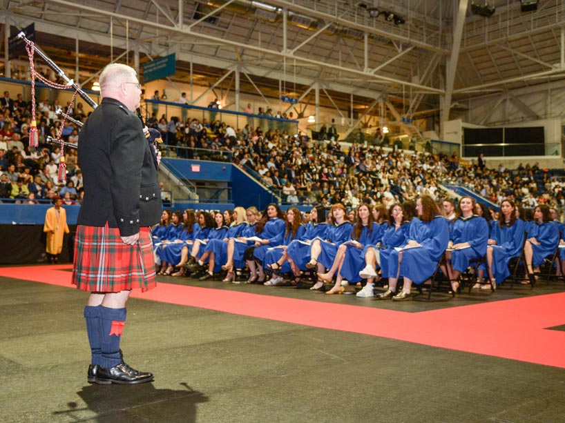 New graduates wearing blue gowns sitting in chairs with a bag piper playing in front