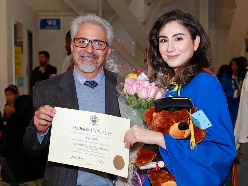 A father holds his daughter's degree, who stands beside him holding flowers and a teddy bear in her blue convocation gown