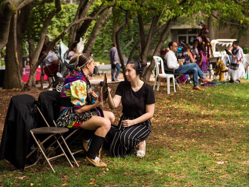 A student (left) holds a mic and wears headphones while interviewing an Indigenous dancer