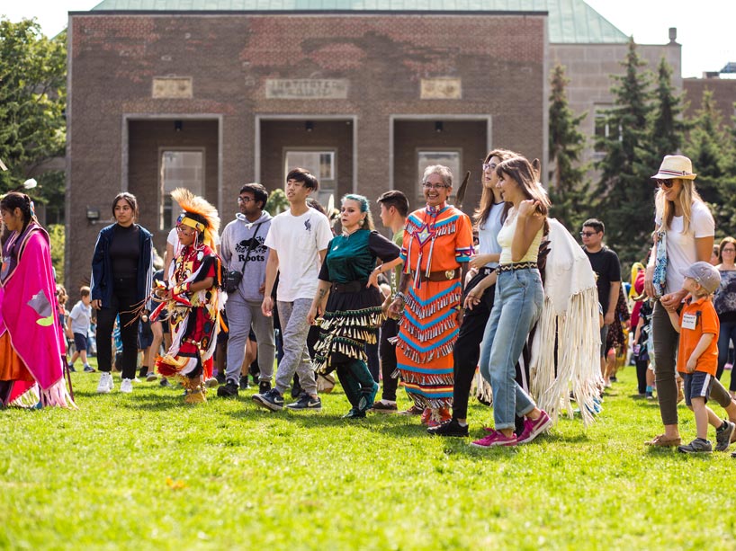 Indigenous dancers wearing regalia lead a group of school children around the quad