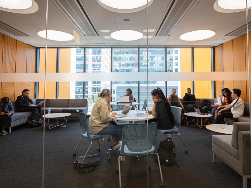 Seen through a window, students are seated at round tables with laptops and on couches in groups.