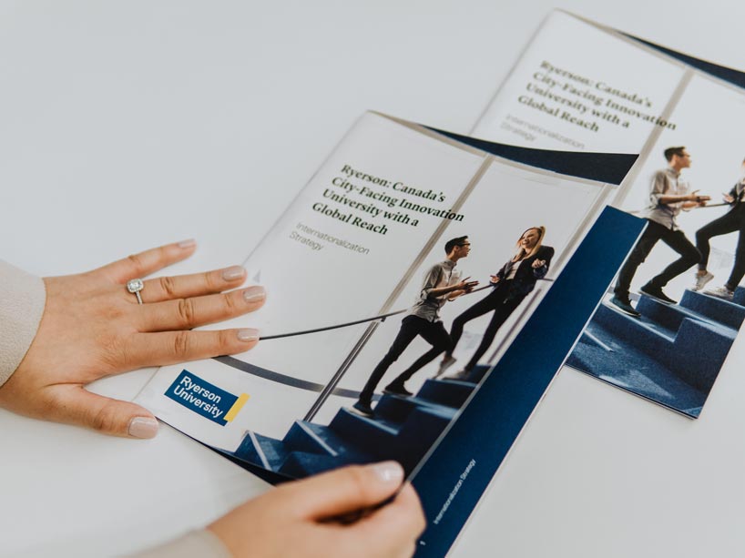 Female hands grasp print copy of Internationalization Strategy, cover is of man and woman talking on staircase, Ryerson logo on bottom left corner