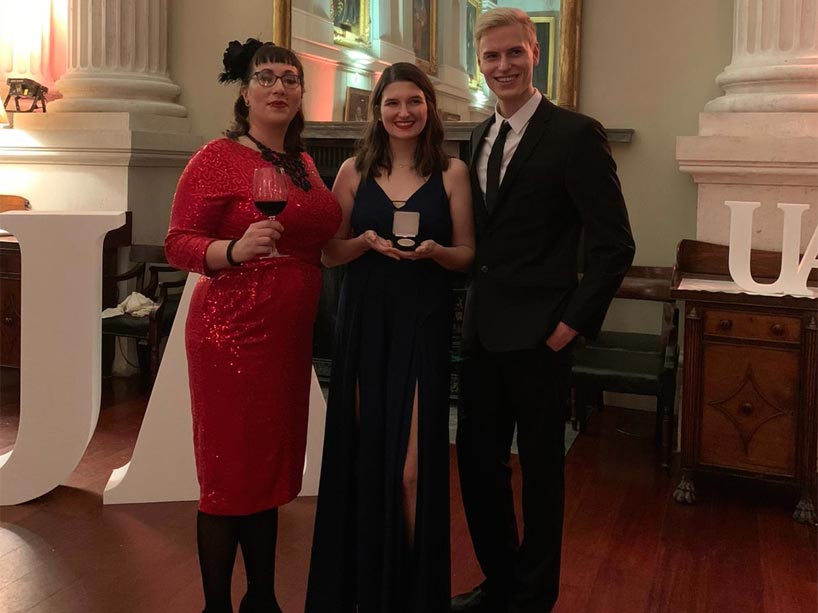 Three award winners, dressed in evening wear, stand. One of them, Laura DeSousa, holds her award.