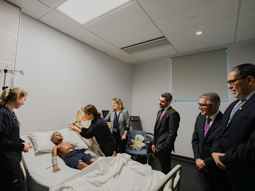 Student stands over simulated hospital bed, practicing on a dummy, as officials watch demonstration