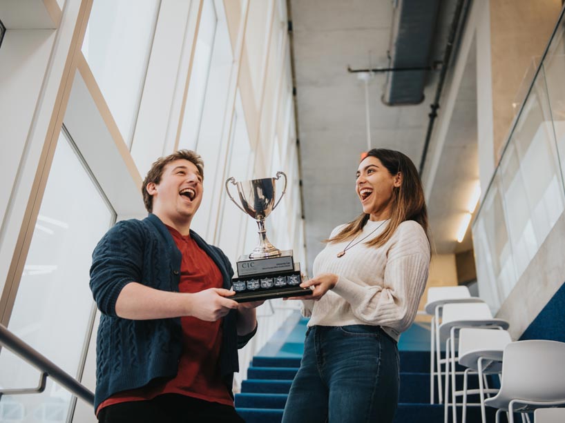 Male and female students smile while both holding silver trophy