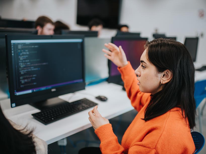 A student sits in front of a computer with her arms raised, working on code with a peer