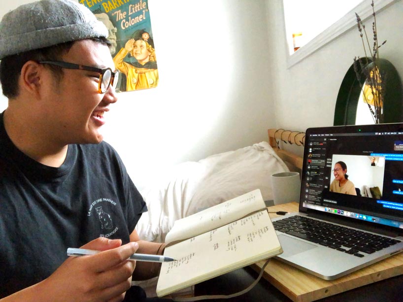 A student holds a pen and notebook while smiling at his laptop during a video chat with a friend