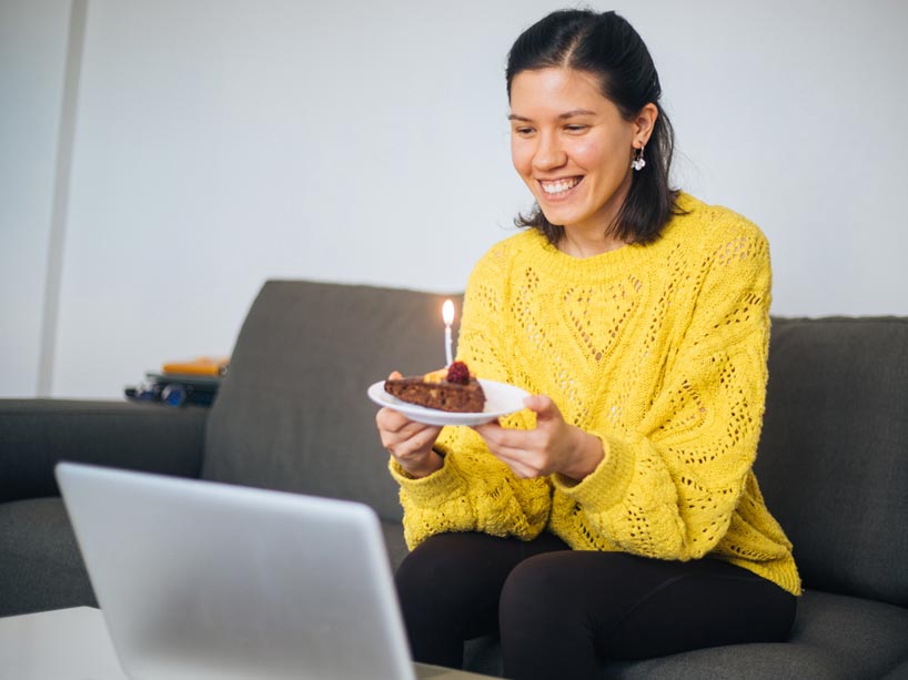 A woman sitting on her couch in front of a laptop holding a plate with a cupcake on it