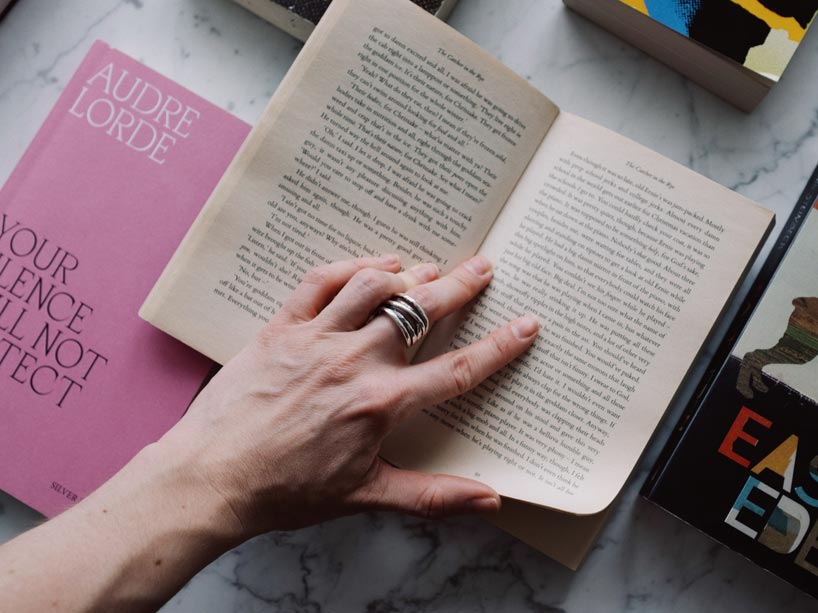 A hand holding open a book with other books around it, laid out on a table