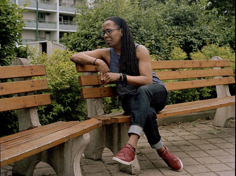 A woman sits on a bench in front of some shrubbery, with a building in the background