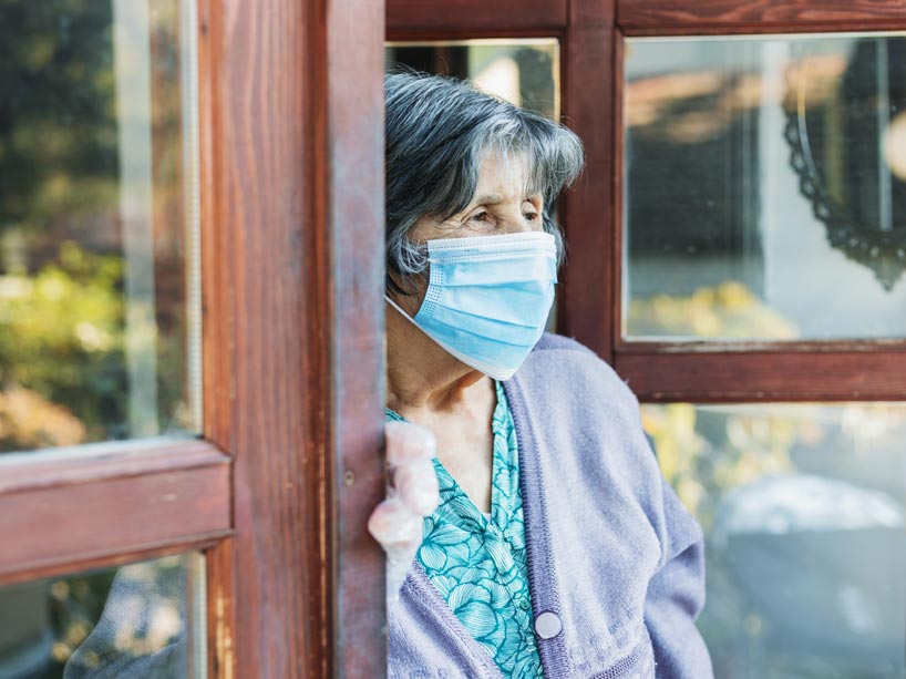 An elderly woman wearing a face mask and protective gloves gazes out from a doorway.