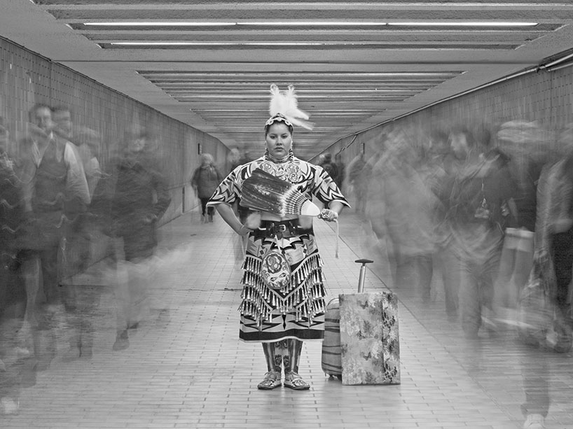 A woman in Indigenous regalia stands between busy commuters