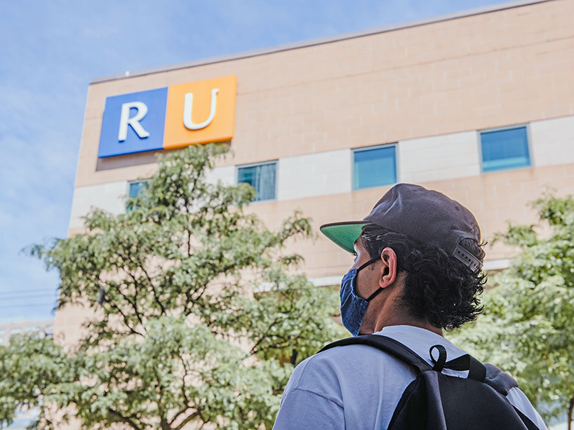 man wearing baseball hat, backpack and mask looks up at campus building in the background