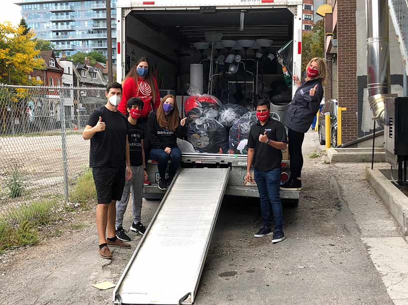 Six people in masks stand at the back of an open truck loaded with furniture and bags of clothing.