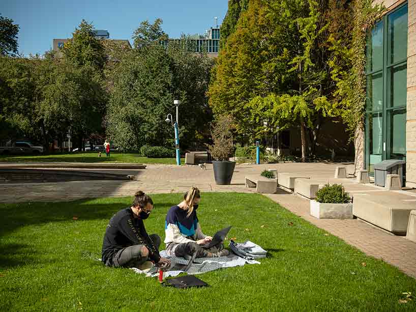 Two students wearing masks sit on the grass in front of Pitman Hall working on laptops