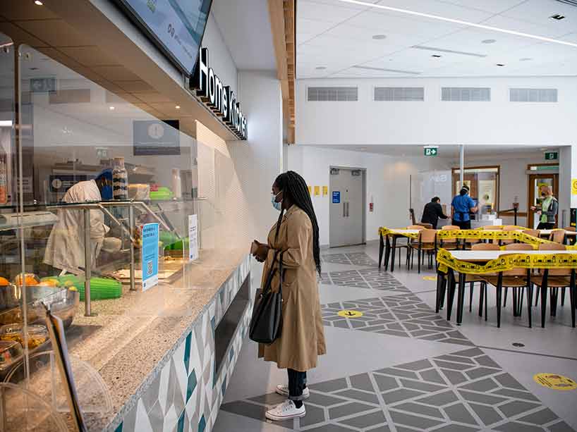 A woman wearing a mask stands at the cafeteria servery. Empty tables are marked with yellow tape. One person stands in the background.