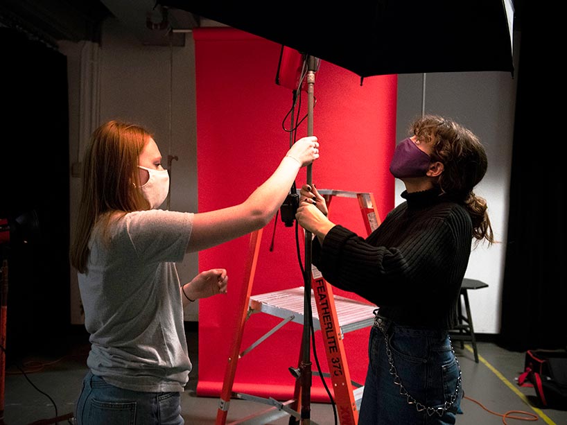 Two students in masks stand and adjust a lighting umbrella in front of an orange backdrop in a photo studio.