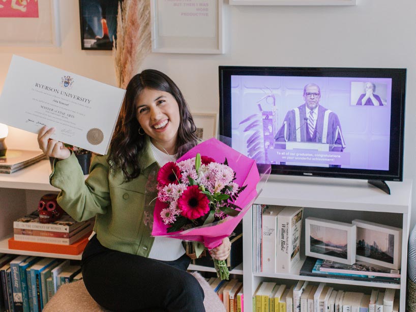 Alia Youssef smiles holding her degree and a bouquet of flowers, with Ryerson’s virtual graduation celebration playing on a screen in the background