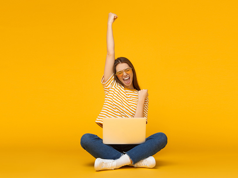 A woman with a raised fist acts excited as she sits with her laptop in front of a yellow background