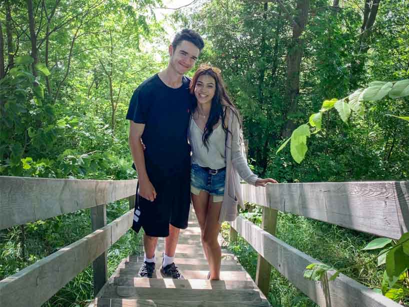 A couple smiles while standing on the stairs outside surrounded by trees.