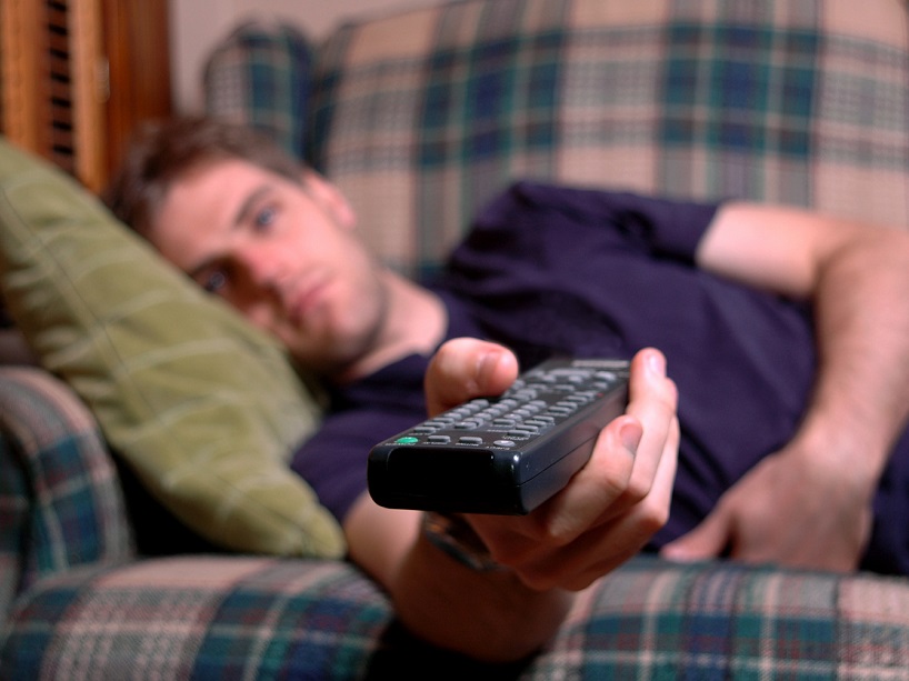 A middle-aged man lies on the couch with a blank expression, TV remote in hand.