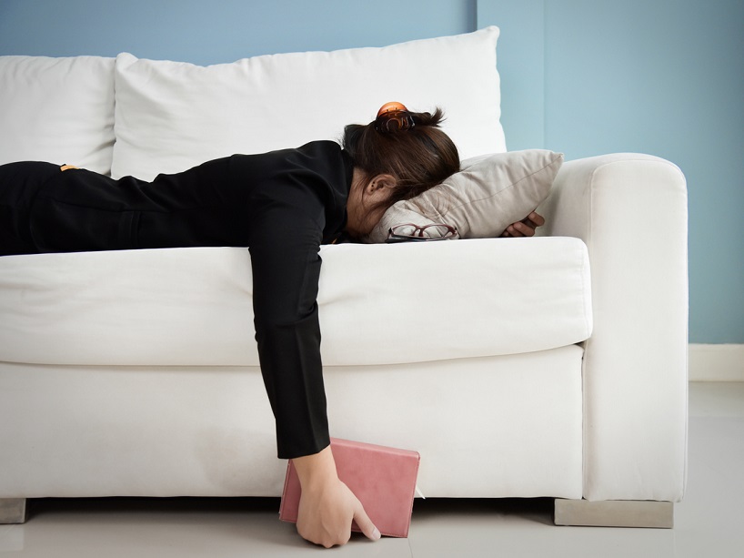 Exhausted woman is collapsed on couch, face down, holding a notebook in her hand.