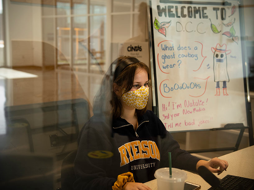 Student in Ryerson sweatshirt and wearing a mask sits behind plexiglass at a front desk in residence.