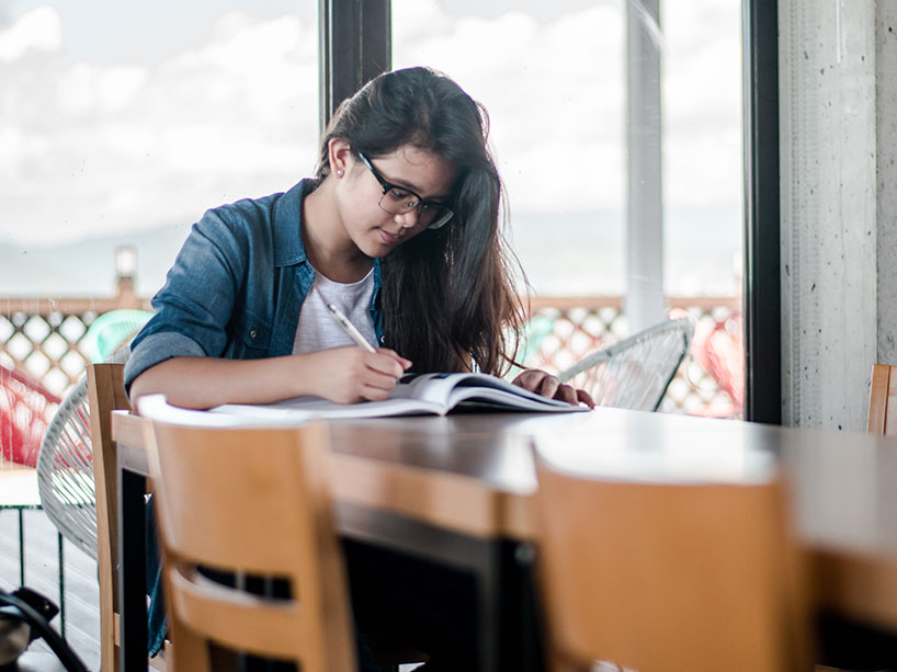 Woman sitting at a table in front of a large window writing into a notebook.