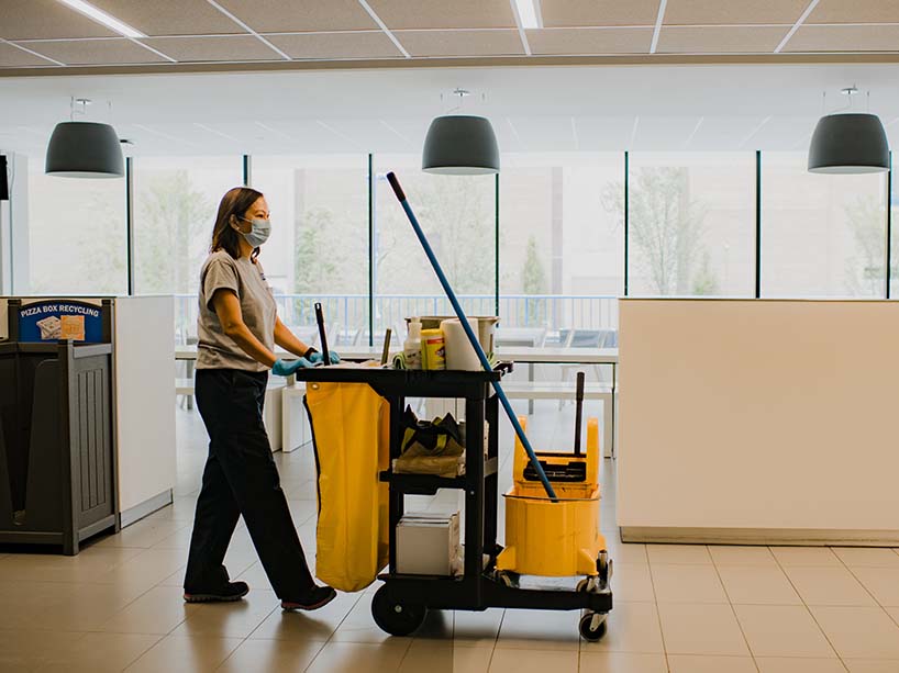 A frontline worker pushing a cart with cleaning supplies down a hallway.