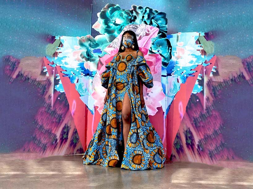 A model in a blue and orange patterned dress and matching face mask walks down a runway, with a bright splash of vibrant colour behind her.