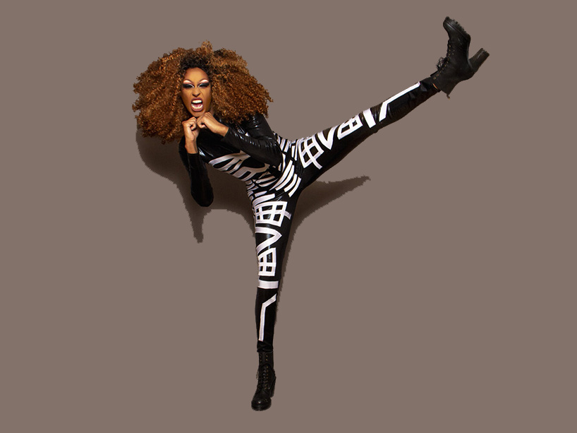 Priyanka, who is wearing a tight black bodysuit with a white design, kicks one leg high in the air, fists raised toward her chin, mouth wide open with an excited expression.