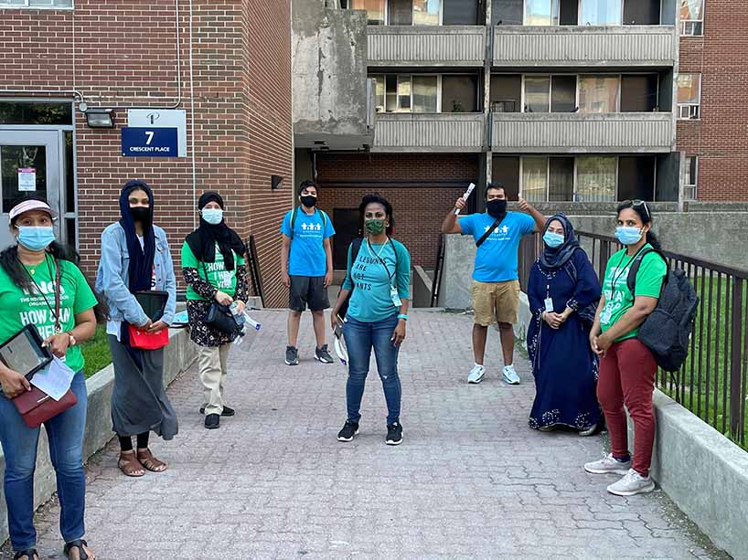 Eight people standing outside of an apartment building wearing masks. 