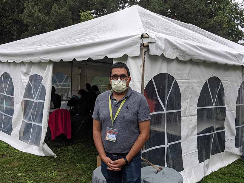 A man wearing a mask and badge standing outside of a white tent outdoors. 