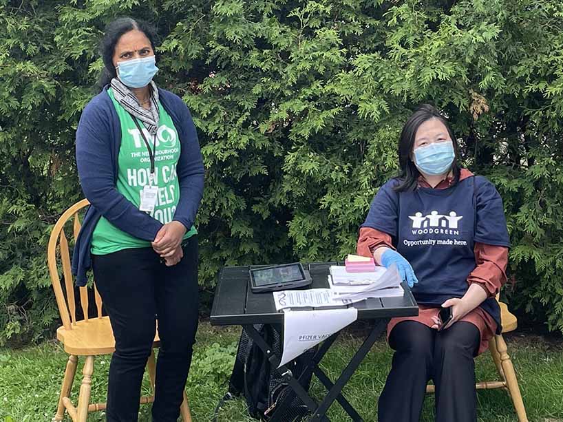 Two women beside a pop-up desk with papers, wearing masks outside. 