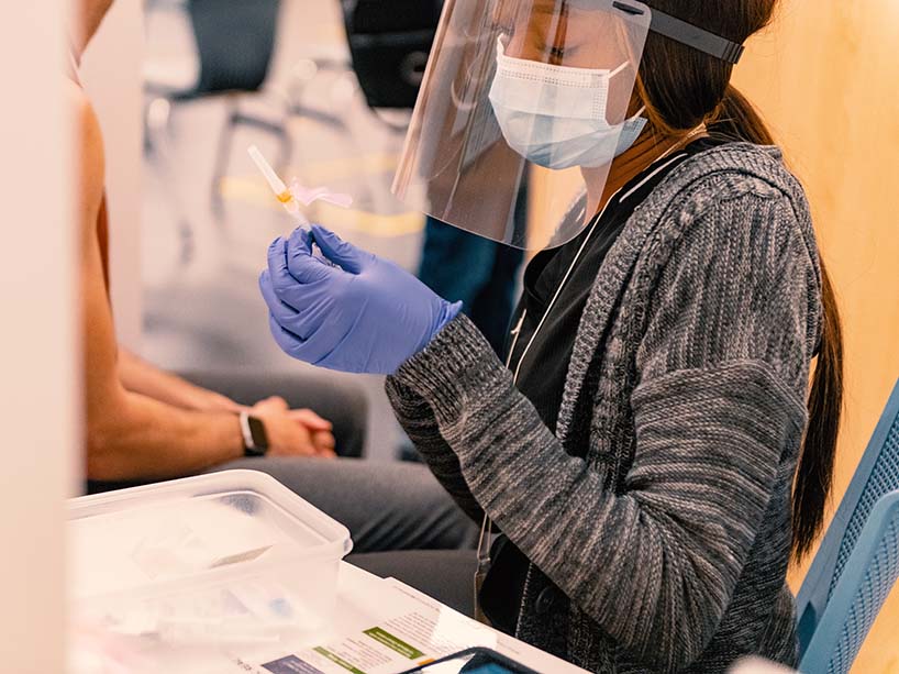 A nurse wearing a mask and face shield unwrapping a syringe.