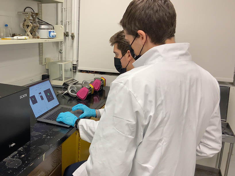 Two men in masks and white coats in a lab working with drug-checking device and laptop.