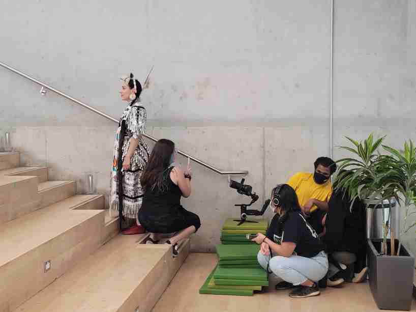 Members of the education week and Pow Wow organizing team are seen in a stairwell filming a dancer in regalia for the Pow Wow video. 