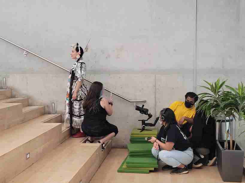 Members of the education week and Pow Wow organizing team are seen in a stairwell filming a dancer in regalia for the Pow Wow video.