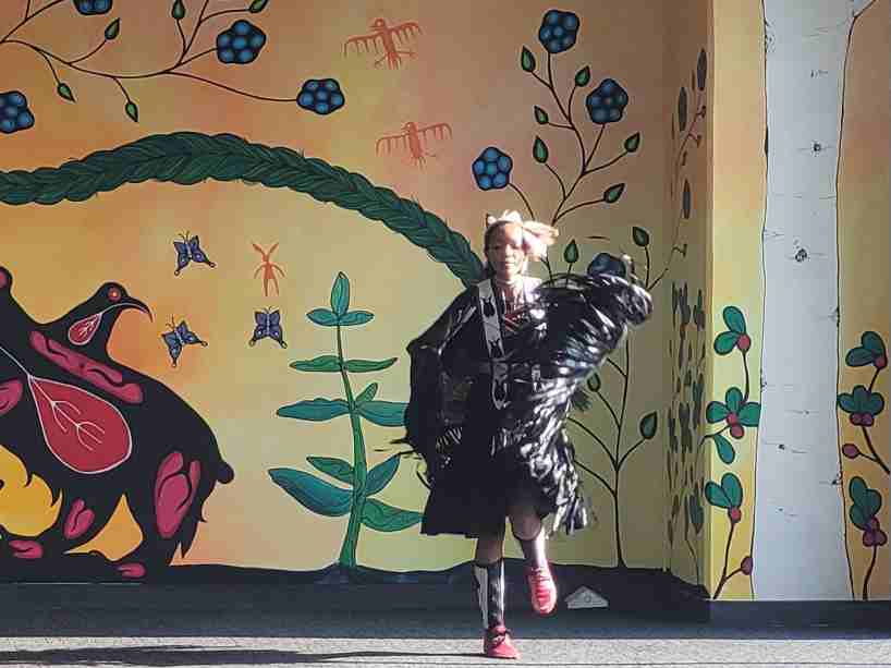 A youth dancer is seen in regalia in front of a vibrant coloured wall during the filming of the 2021 Pow Wow.